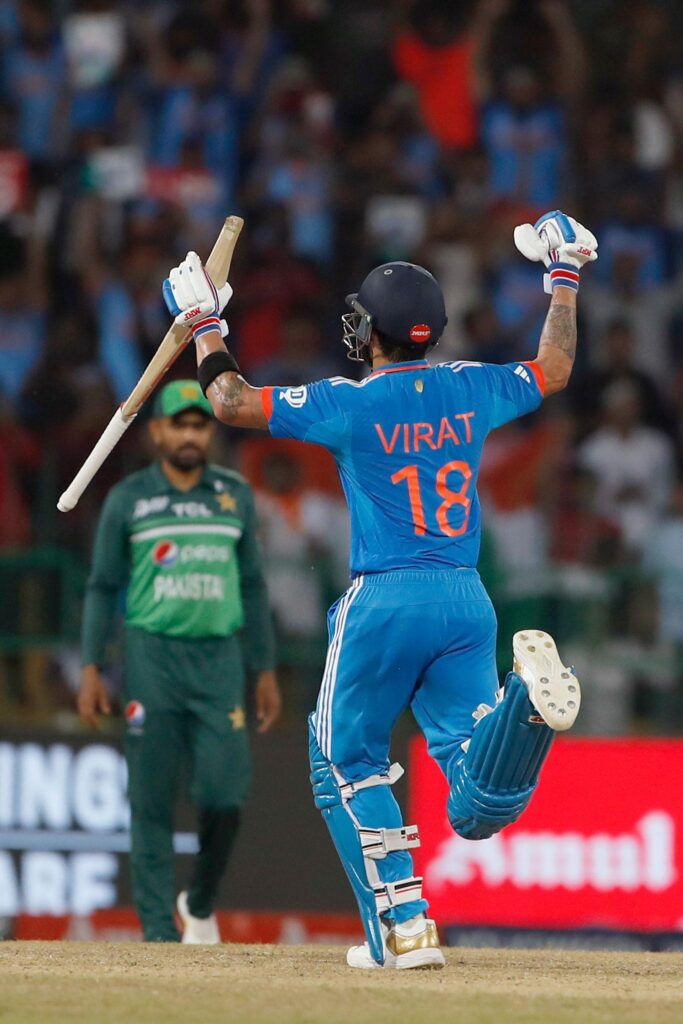 Virat Kohli in blue India jersey raising his bat in celebration during a high-voltage India vs Pakistan cricket rivalry match, with a Pakistan player in the background.india 